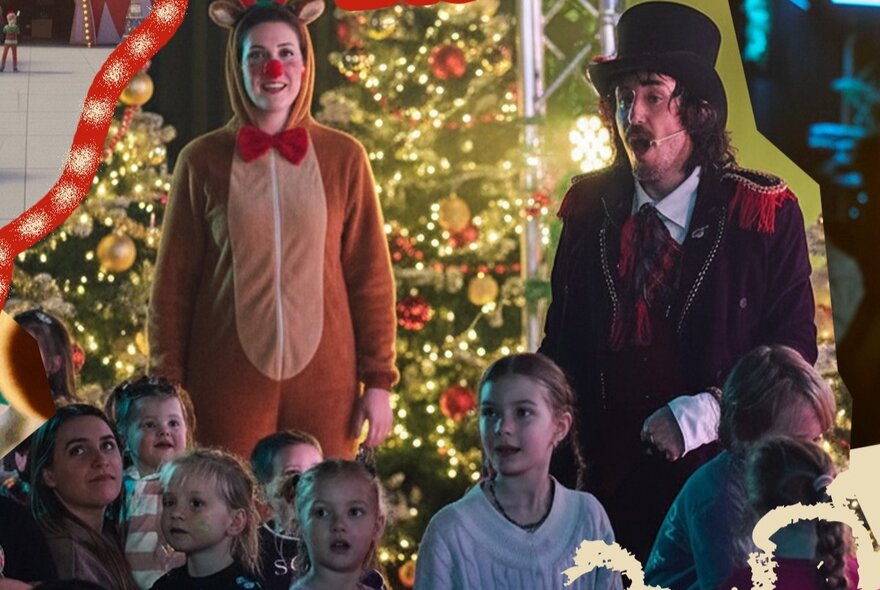 A group of young children at an outdoor performance, in the background is a large Christmas tree, a man in a top hat and jacket singing and a person dressed as a reindeer.