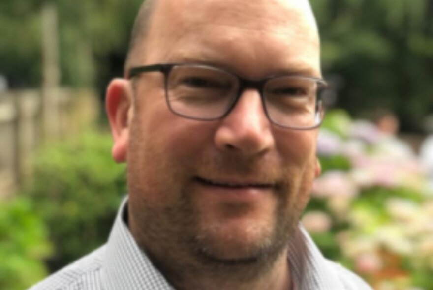 Portrait of a man wearing glasses, in front of a blurred floral garden background.