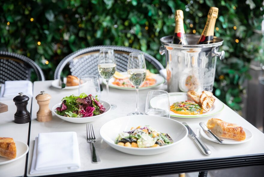 An outdoor restaurant table set for a meal, featuring several white plates of food including a leafy green salad and pasta dishes and two wine glasses.