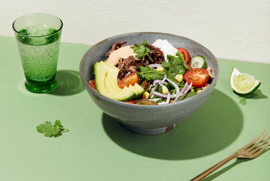 Salad bowl, green glass of water, coriander leaf and quarter of lime on a green surface.