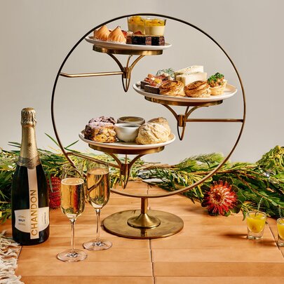 A three-tier stand of high tea food items, a bottle of sparkling and two glasses, arranged on a wooden table with bunches of native flora in the background.