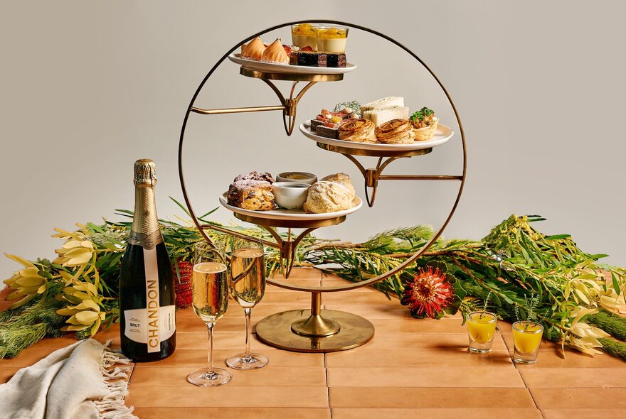 A three-tier stand of high tea food items, a bottle of sparkling and two glasses, arranged on a wooden table with bunches of native flora in the background.