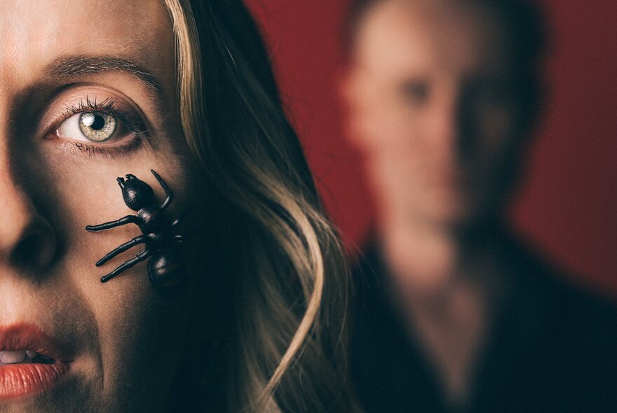 Half of a woman's face with a large plastic ant resting on her cheek, a man looking on in the background though completely blurred.