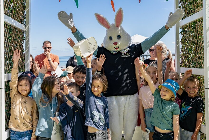 Children gathered around a person in an Easter bunny costume during an outdoor event under a blue sky.