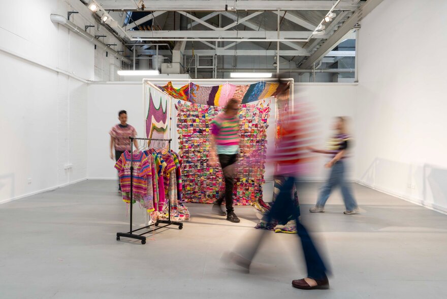 People in motion in a white exhibition space, walking around a centrally positioned installation.