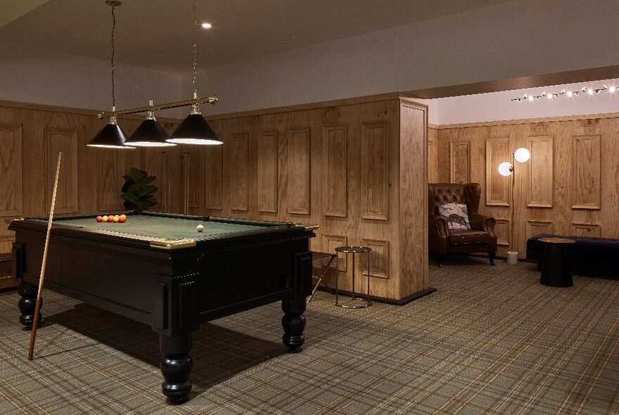 A pool table in a billiard room with patterned carpet and timber panelling on the walls; a small sitting area with leather chesterfield armchair behind.