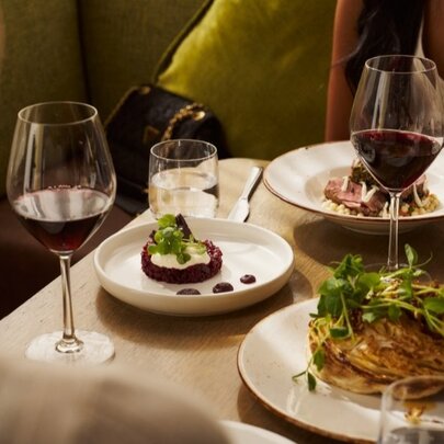 Plates of food on a restaurant table, with two glasses of red wine and two people seated opposite each other. 
