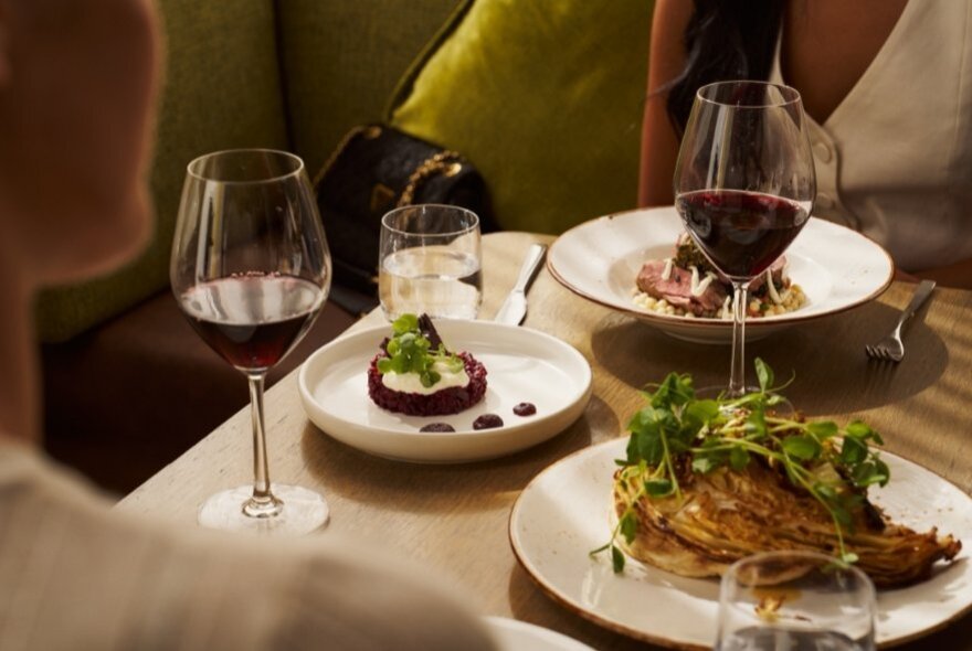 Plates of food on a restaurant table, with two glasses of red wine and two people seated opposite each other. 