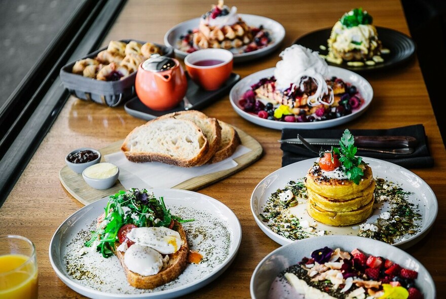 Window bench laden with breakfast dishes: eggs on toast, pancakes, waffles, toast and orange juice.