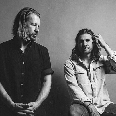 Ragged-edge photo from contact sheet, two men, one with hands clasped in front of him, the other seated brushing his hair from his face.
