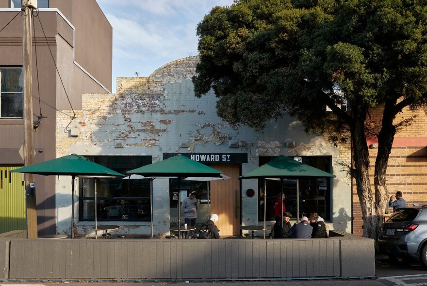 Exterior of Code Black Cafe showing three green market umbrellas, seating outside on the footpath and a large tree in front of the cafe