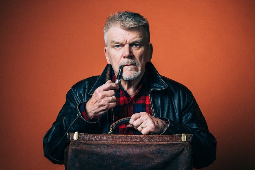 A man looking directly at the camera, holding a pipe to his mouth and an old-fashioned leather bag, against an orange background.