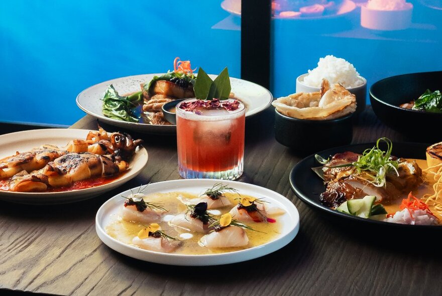 An array of delicious-looking traditional Thai dishes displayed on a wooden table, with a short cocktail in the middle.