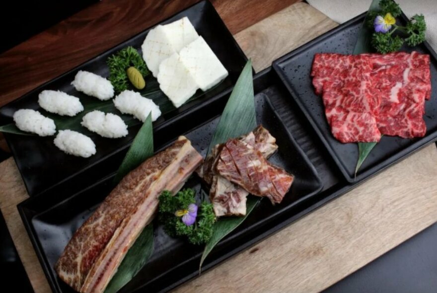 Cuts of meat and sushi rice on black plates.