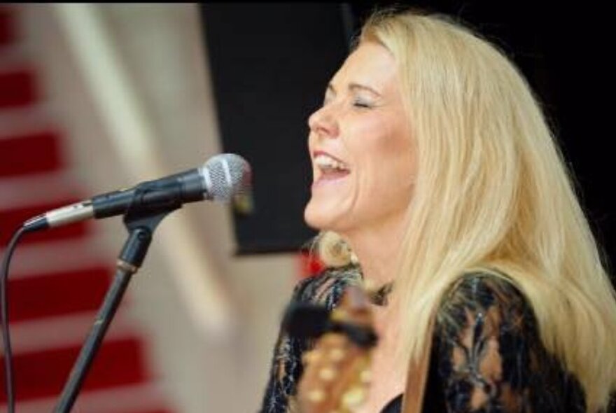 Blonde performer singing into a microphone with her eyes closed, wearing a black lace dress.