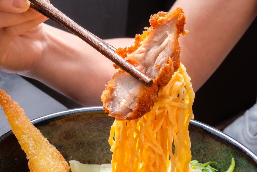 A hand using chopsticks that hold fried chicken and yellow noodles.
