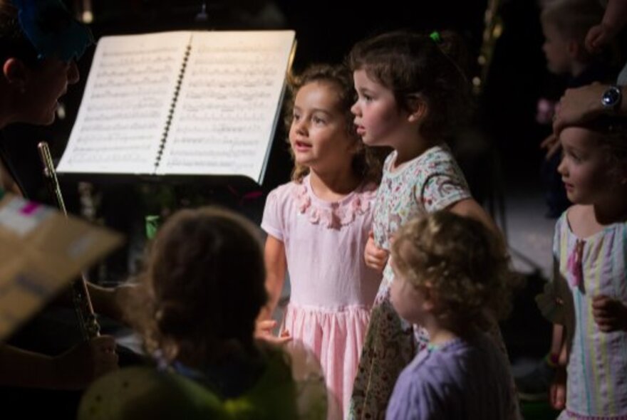 Young children listening to music being performed, a music stand with open sheet music is visible to the left of the frame.