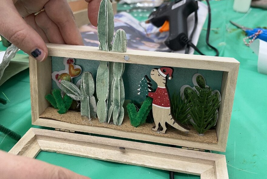 A hand showing a diorama featuring a festive dinosaur.