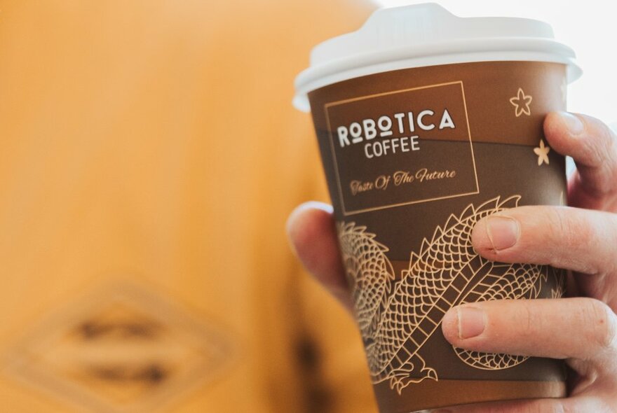 A hand holding a brown takeaway coffee cup in front of an orange background.