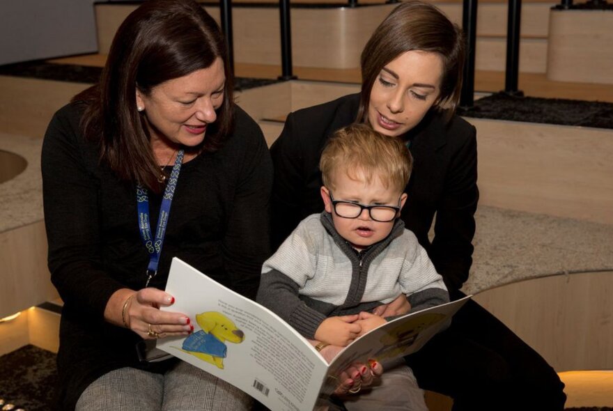 Two adults and a young child looking at a picture book, in a library setting.
