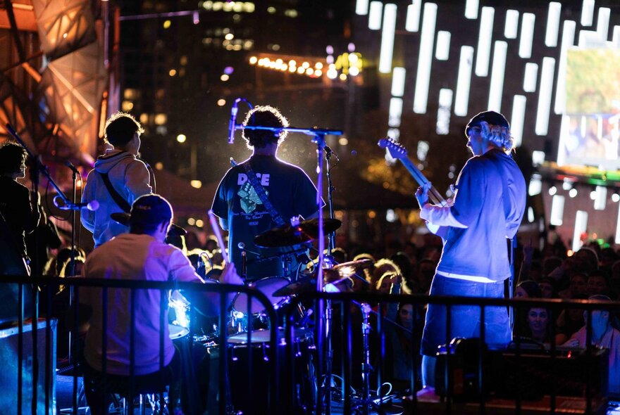 A young band on stage at Fed Square at night, seen from behind, with three guitarists and a drummer.