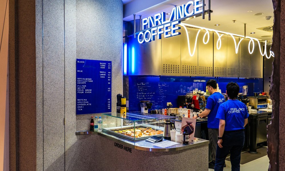 Two baristas in a tiny cafe with bright blue and silver walls and a neon light. 