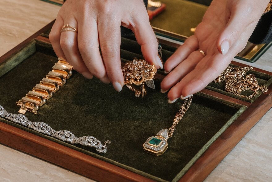 A woman's hands holding a gold broach over a velvet-lined display case with other gold jewellery pieces in it.