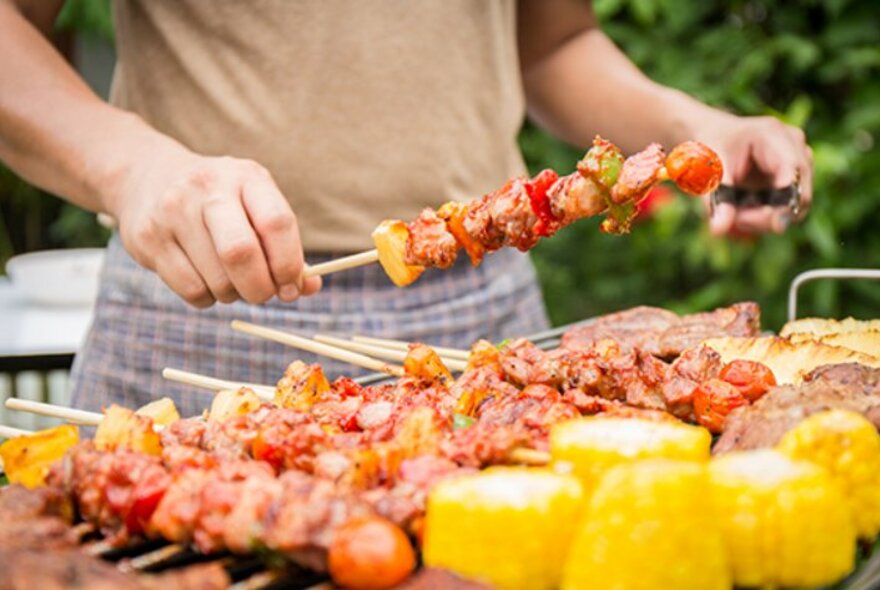 A person cooking food on a barbeque, picking up a skewer of meat cubes and vegetables.