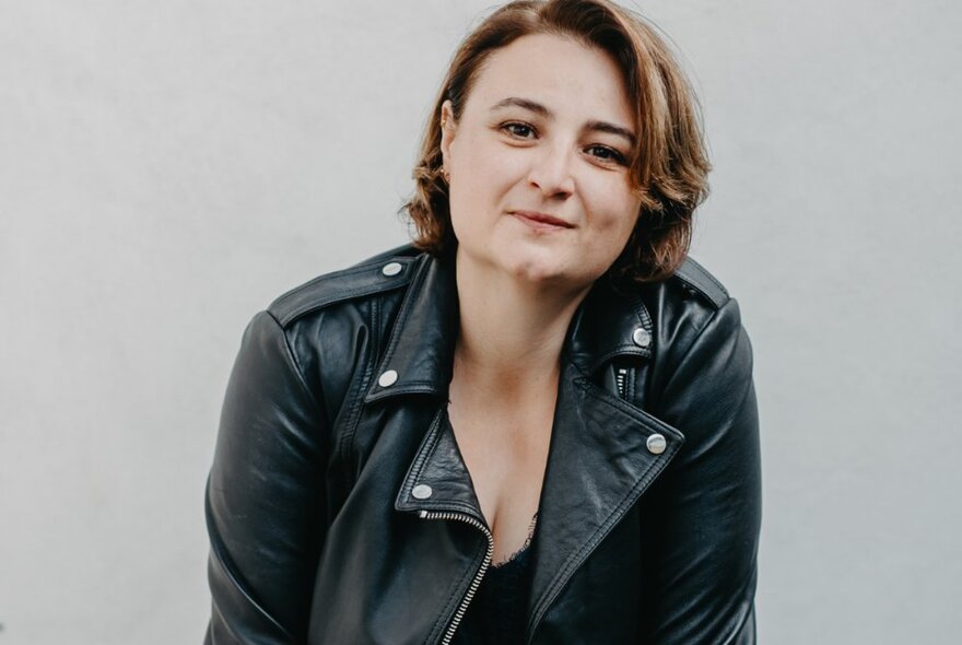 Portrait of a subtly smiling woman wearing a leather jacket, in front of a light grey background.