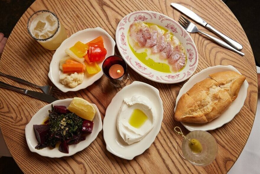 Looking down at a table at several dishes of Greek food on a wooden table, including a creamy dip, pickled vegetables, crusty bread.