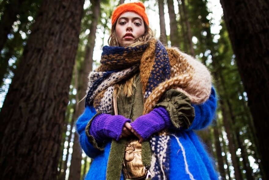 Looking from below at a woman in a forest with tall, straight trees, wearing a red beanie and woollen knits and scarves.