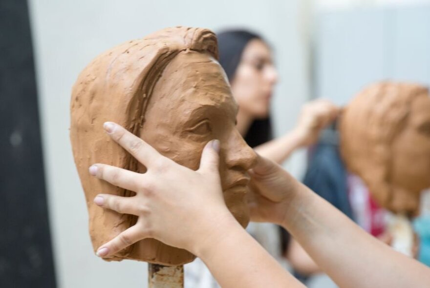 Hands working on a sculpture of a head using clay. 
