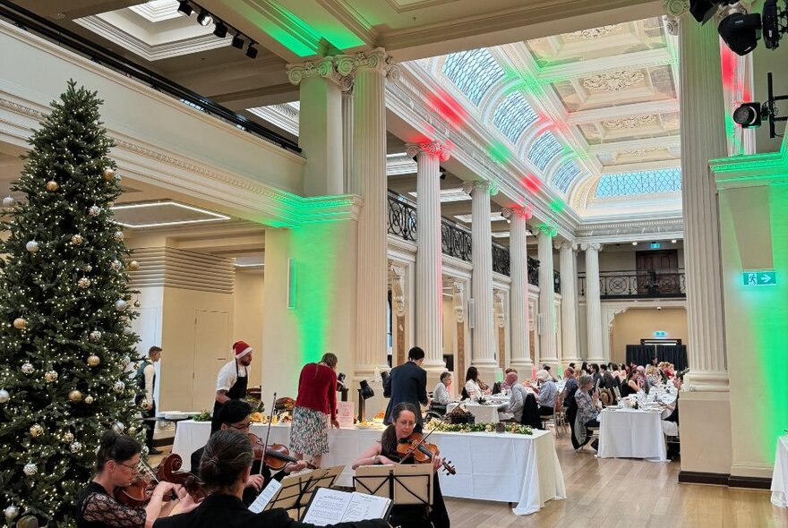 A room in the State Library Victoria set for Christmas dining, with a Christmas tree in the corner, a string quartet and coloured lights.