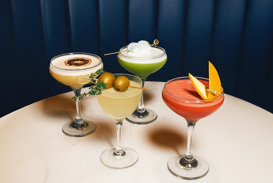 Four differently coloured cocktails in martini glasses on a white table.