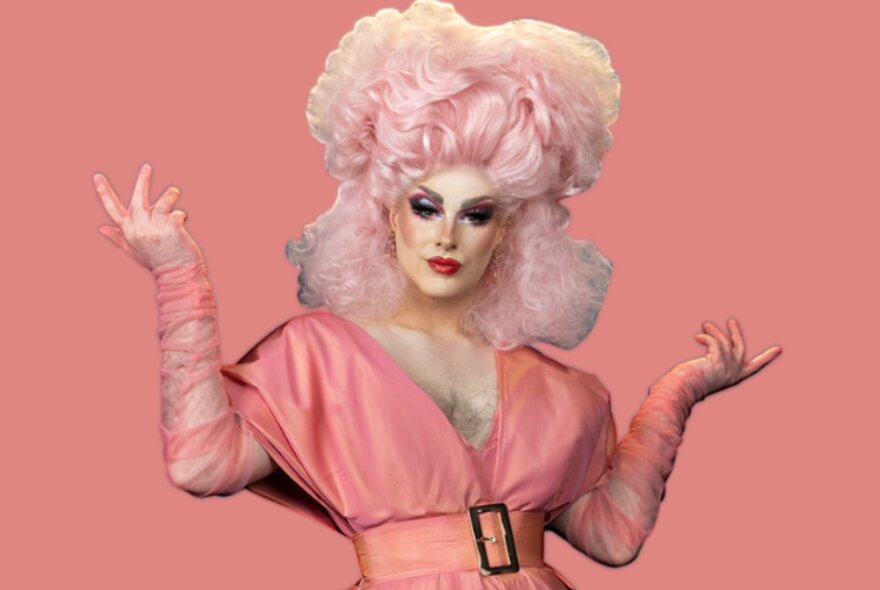 A drag queen with hands arranged upwards wearing an 80s-style apricot dress with a side belt and a powder pink wig.