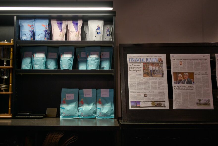 A newspaper display, and packaged coffee for sale on shelves inside a cafe.