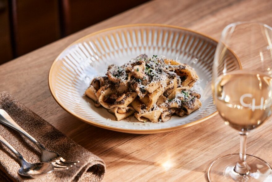 Bowl of pasta on wooden table, glass of white wine to right, front.