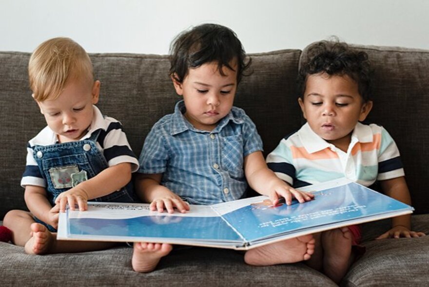 Three toddlers seated together on a couch, a large picture book resting open across their legs.