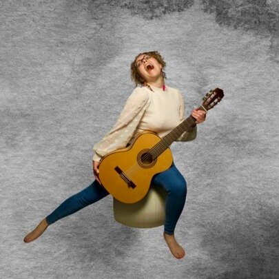 Woman holding an acoustic guitar, seated on a ball amid clouds, her head back singing.