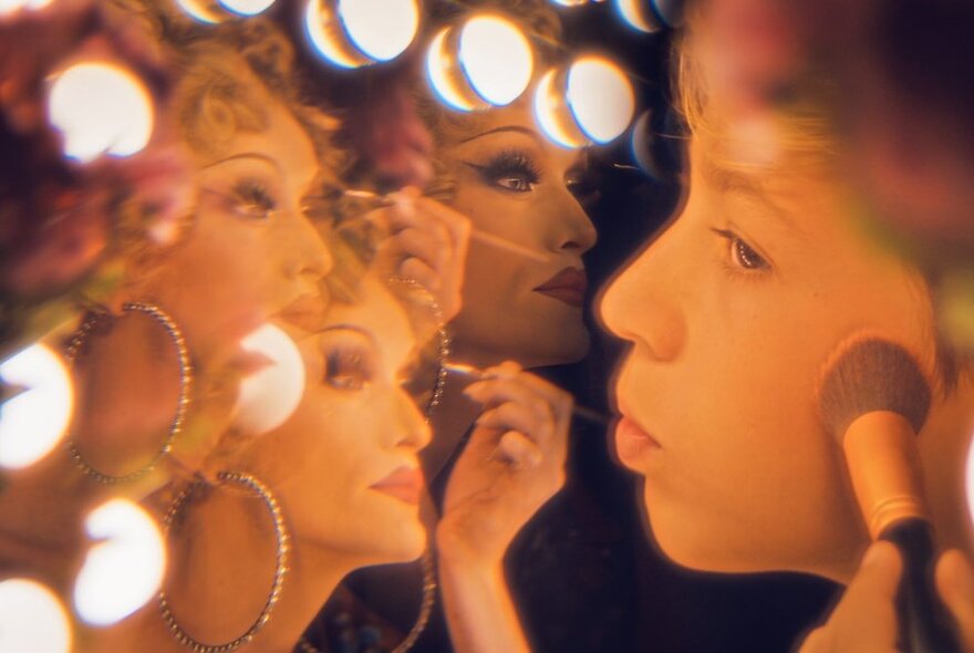 A still from a film of a young teenage boy brushing makeup onto their cheek looking into a mirror that reflects multiple images of a drag artist in full makeup.