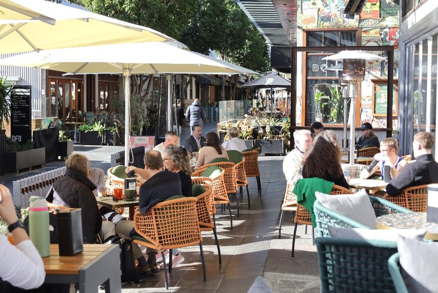 People seated at outdoor cafe tables under umbrellas in dappled sunlight.