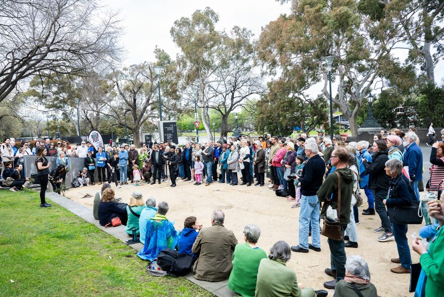 Large group standing listening to a speaker under winter trees in a park setting.