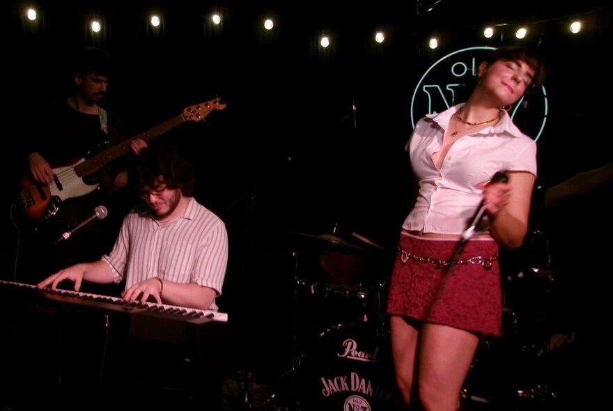 Duo performing on stage under lights, female singer wearing short skirt and blouse, holding microphone; male keyboard player.