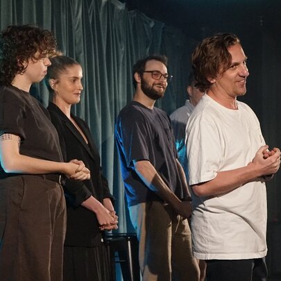 Performers standing in a row on a stage in front of a curtain, with one performer standing slightly in front of the others.