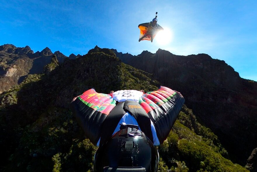 Two people flying through the sky with a mountain behind them, wearing body parachute wings and helmets.