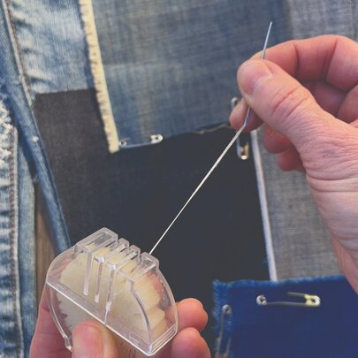 A person's hands holding sewing implements with patches of denim secured with safety pins behind.