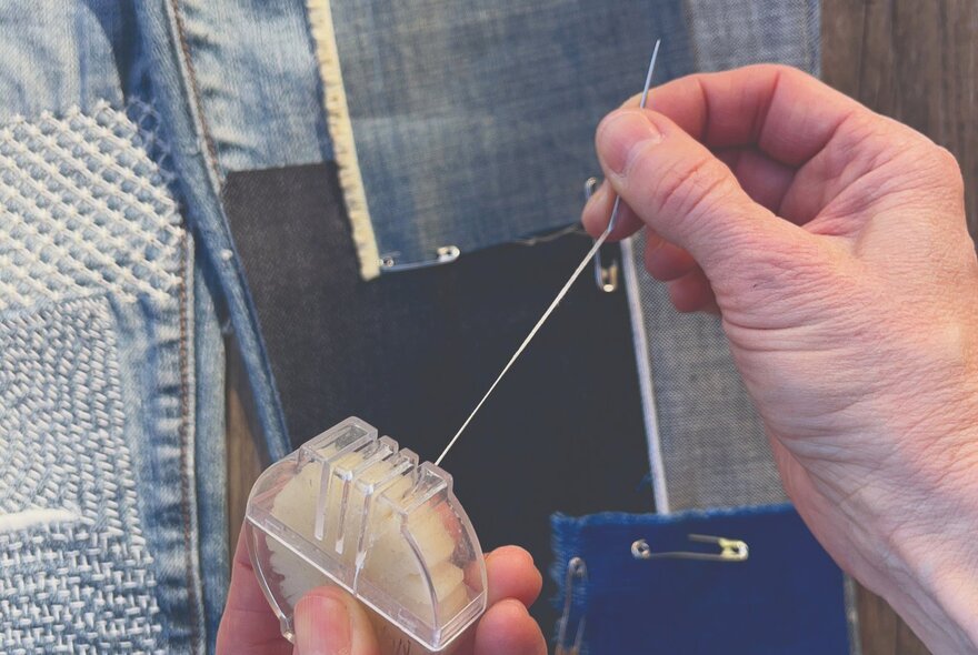 A person's hands holding sewing implements with patches of denim secured with safety pins behind.