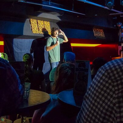 People seated in a dark bar venue watching a stand-up comic performing on stage, holding a microphone.