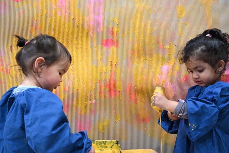 Two little girls in blue art smocks squeezing paint next to wall that has been daubed and splattered with yellow and pink paint. 