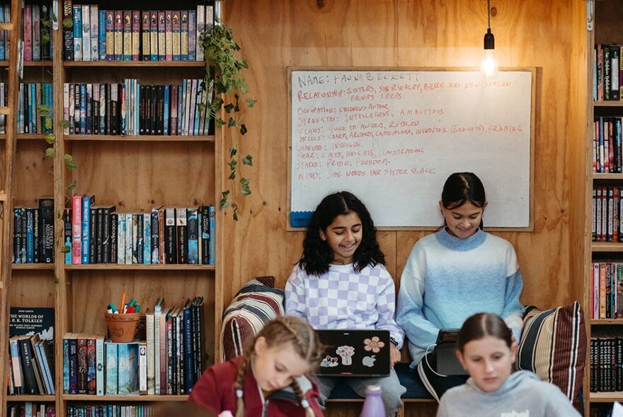 Two young teens writing on their laptops, seated on a couch between two bookcases in a room, two other teenagers seated at a table in front of them.